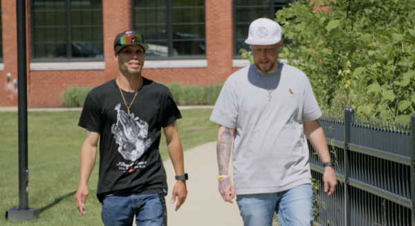 Two men walking on a sidewalk beside a fence, one wearing a black shirt and cap, the other in a gray shirt and white hat. A red brick building is in the background.